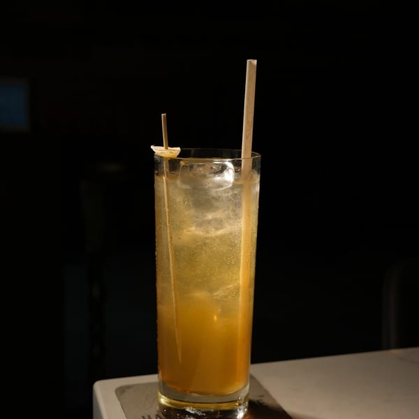 Tall glass of iced amber cocktail with a straw and lemon garnish on a coaster.