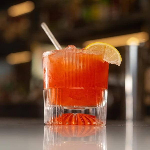 A vibrant red cocktail with ice, a straw, and a lemon slice in a textured glass on a reflective surface.