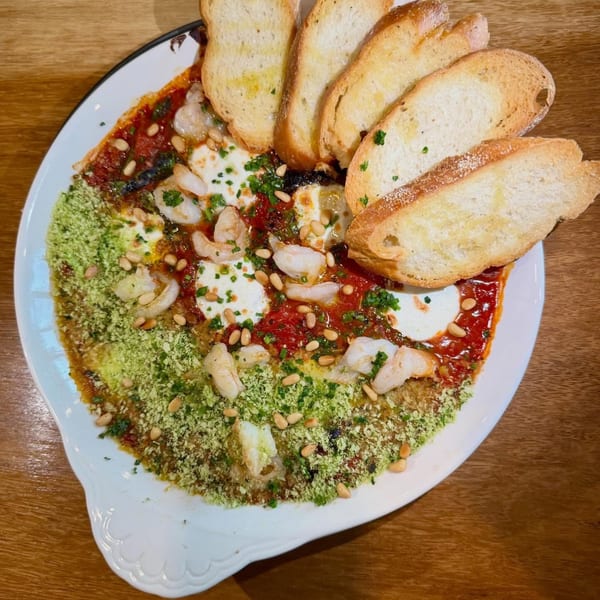 A white platter of baked eggs with tomato sauce, pesto, shrimp, pine nuts, and toasted bread.