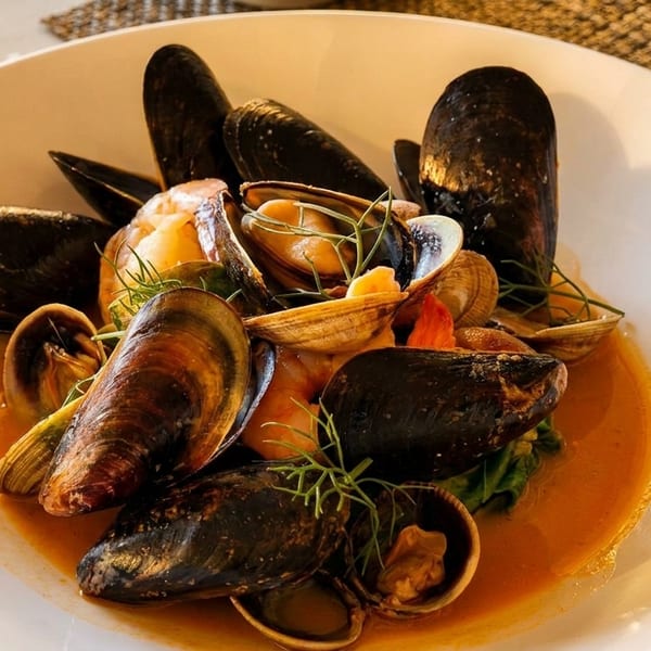 A white bowl filled with mussels, clams, and shrimp in a rich orange broth, garnished with dill.