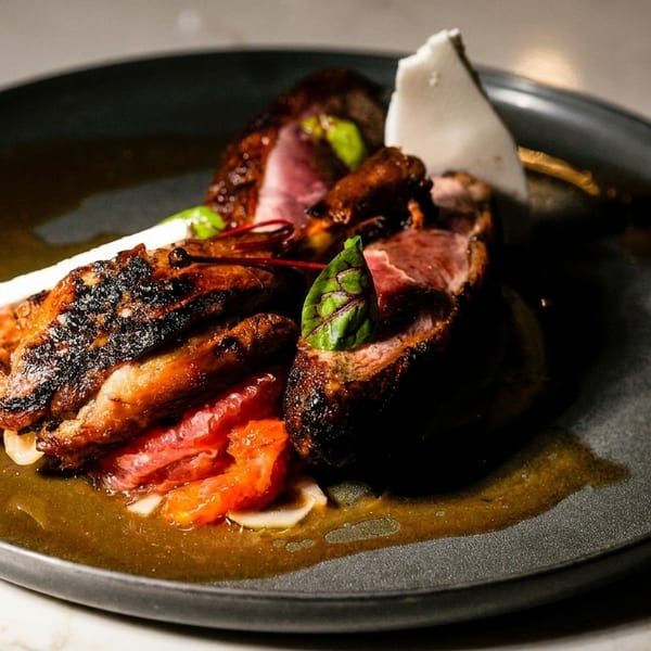 Grilled meat with a rich sauce, red citrus segments, and white garnish on a dark plate.