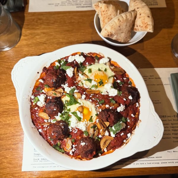A white dish of shakshuka with meatballs, two fried eggs, feta, and fresh herbs, served with pita.