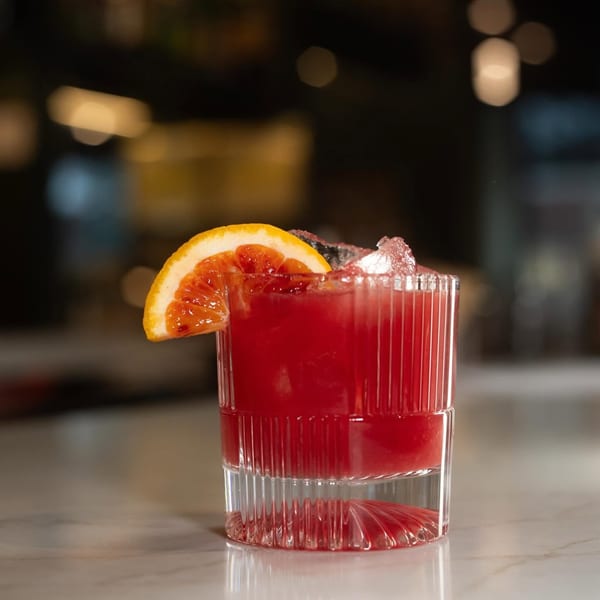 A vibrant red cocktail with ice and a blood orange slice in a textured glass on a marble bar.