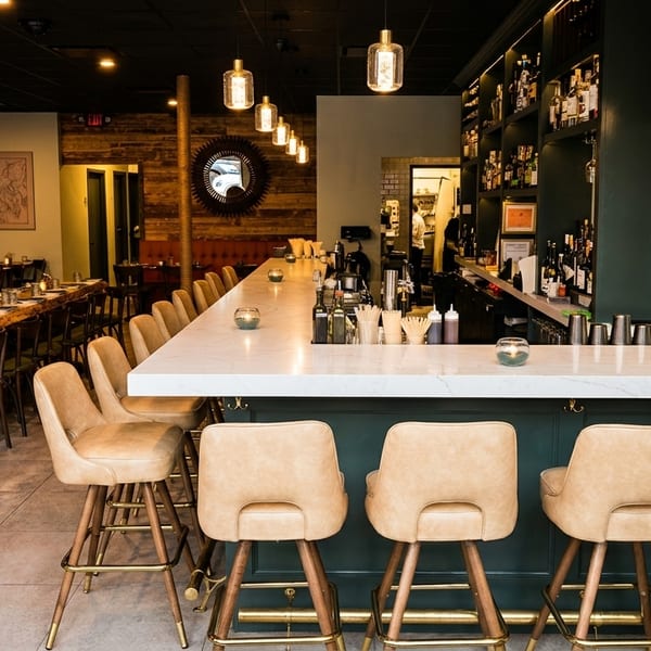 Elegant restaurant interior with a long bar, bar stools, and dining tables with banquette seating.