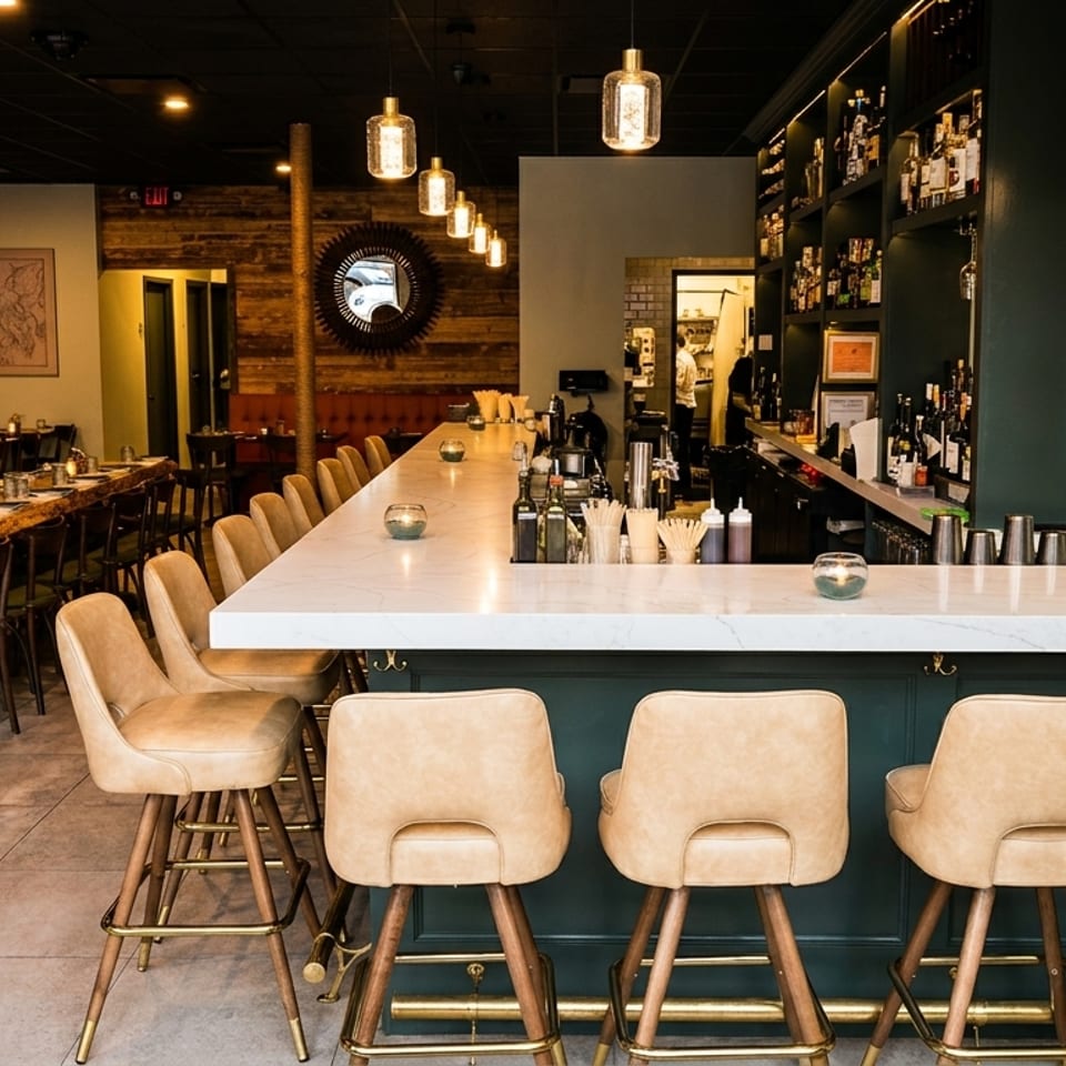 An empty restaurant interior with a long bar, bar stools, and dining tables with banquette seating.
