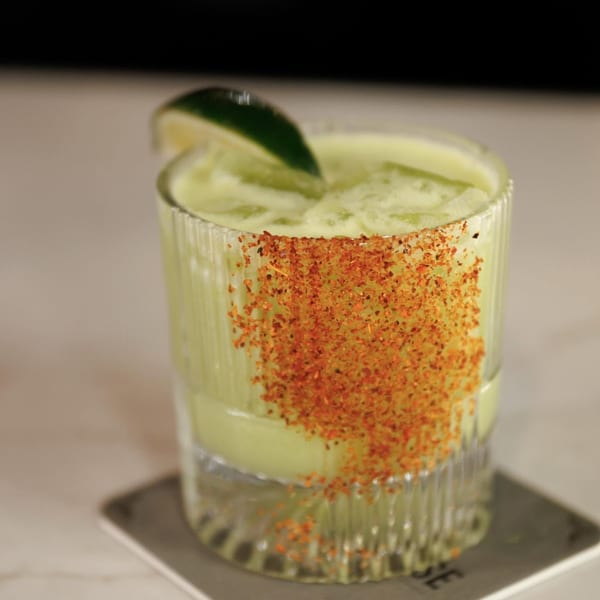 Green cocktail in a textured glass, rimmed with chili flakes and garnished with a lime wedge.